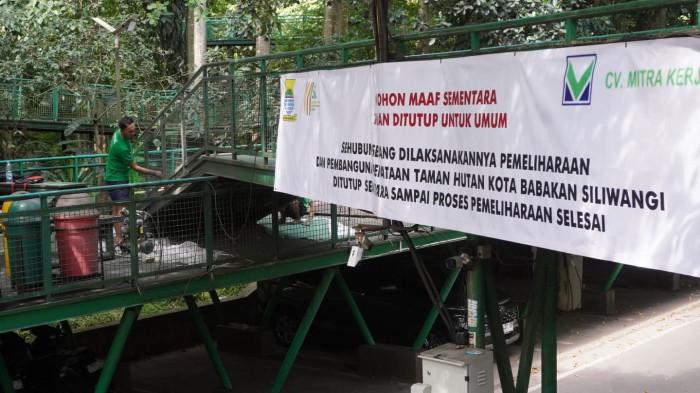 Seorang pekerja melakukan pengecatan ulang pagar besi di samping spanduk berisi pengumuman penutupan sementara taman hutan kota Babakan Siliwangi (Baksil) untuk umum yang dipasang membentang di pagar skywalk kawasan hutan kota Baksil, Kota Bandung, Jawa Barat, Kamis (16/10/2025).