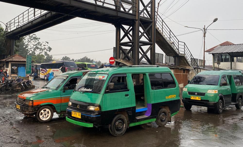Ilustrasi angkutan kota (angkot) berwarna-warni di jalanan Kota Bandung yang ramai, dengan latar belakang gedung-gedung perkotaan dan langit senja.