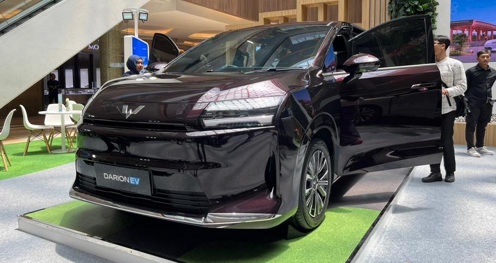 Sebuah mobil Wuling Darion berwarna cerah sedang diproduksi di jalur perakitan pabrik modern di Cikarang, Jawa Barat, dengan beberapa teknisi lokal mengawasi prosesnya. Latar belakang menunjukkan komponen otomotif dan robot industri, melambangkan produksi lokal dan teknologi canggih.