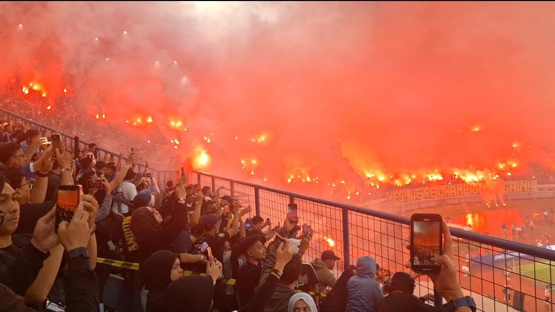 Sebuah foto tribun penonton di stadion sepak bola yang penuh sesak dengan suporter mengenakan syal dan bendera Persib Bandung, menunjukkan antusiasme yang tinggi saat pertandingan kandang.