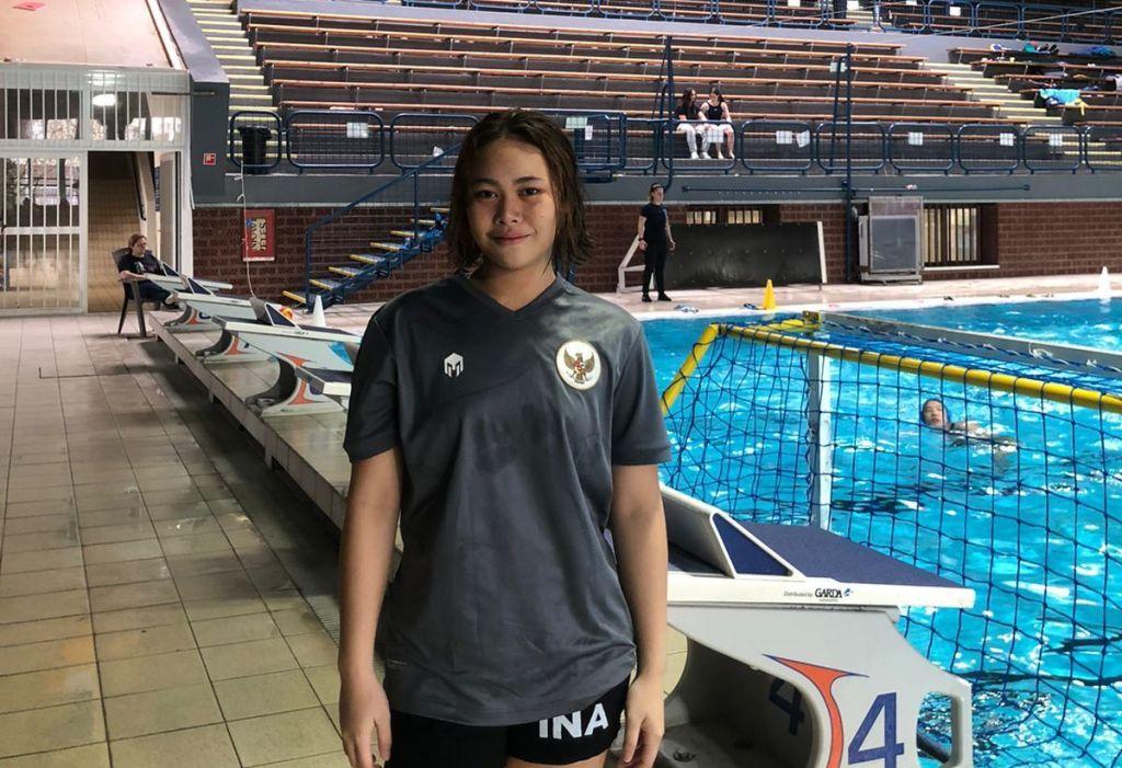 Seorang perenang Indonesia berpose dengan medali emas di lehernya setelah memenangkan kompetisi di SEA Games, dengan latar belakang kolam renang dan bendera Indonesia.