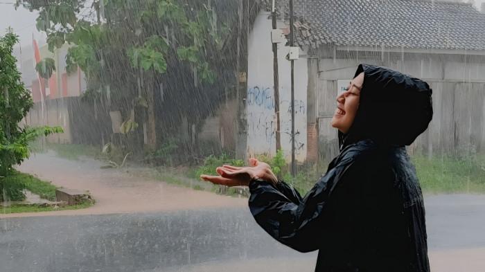 HUJAN LEBAT- Seorang wanita yang sedang menggunakan mantel hujan tampak tersenyum riang menampung cucuran hujan yang jatuh dari langit.