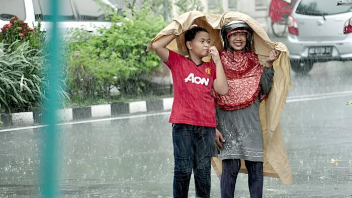 HUJAN SEDANG- Ibu dan anak tampak berjalan menembus hujan sedang yang melanda kawasan tempat tinggalnya. Mereka menghindari hujan dengan mantel seadanya.