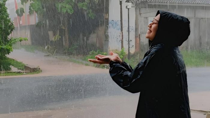 HUJAN SORE HARI- Ilustrasi seorang wanita menggunakan mantel sedang menikmati hujan yang terjadi pada sore hari.