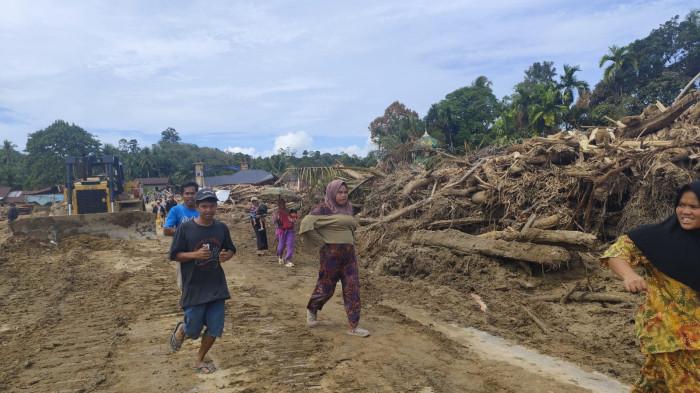 BERLARI - Warga belari saat melintasi jalan bertumpuk gelondongan kayu di Garoga, Batangtoru, Tapsel (4/12/2025). Kondisi di desa ini muncul sebuah aliran sungai baru yang menerjanga rumah warga saat banjir bandang.