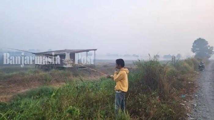 KARHUTLA DI KALSEL - Pemancing di Peramuan menutup hidung ditengah kabut asap, Jumat (21/8/2026). Menurut warga kabut asap tak sepekat dua hari ini