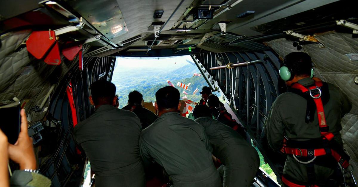 Pesawat C130 J Super Hercules TNI Angkatan Udara sedang melakukan airdrop (penerjunan) bundle-bundle bantuan logistik menggunakan parasut di atas area pedesaan yang sebagian terendam banjir di Kabupaten Aceh Gayo. Langit cerah, dan terlihat beberapa parasut berwarna-warni yang membawa kotak bantuan turun ke daratan.